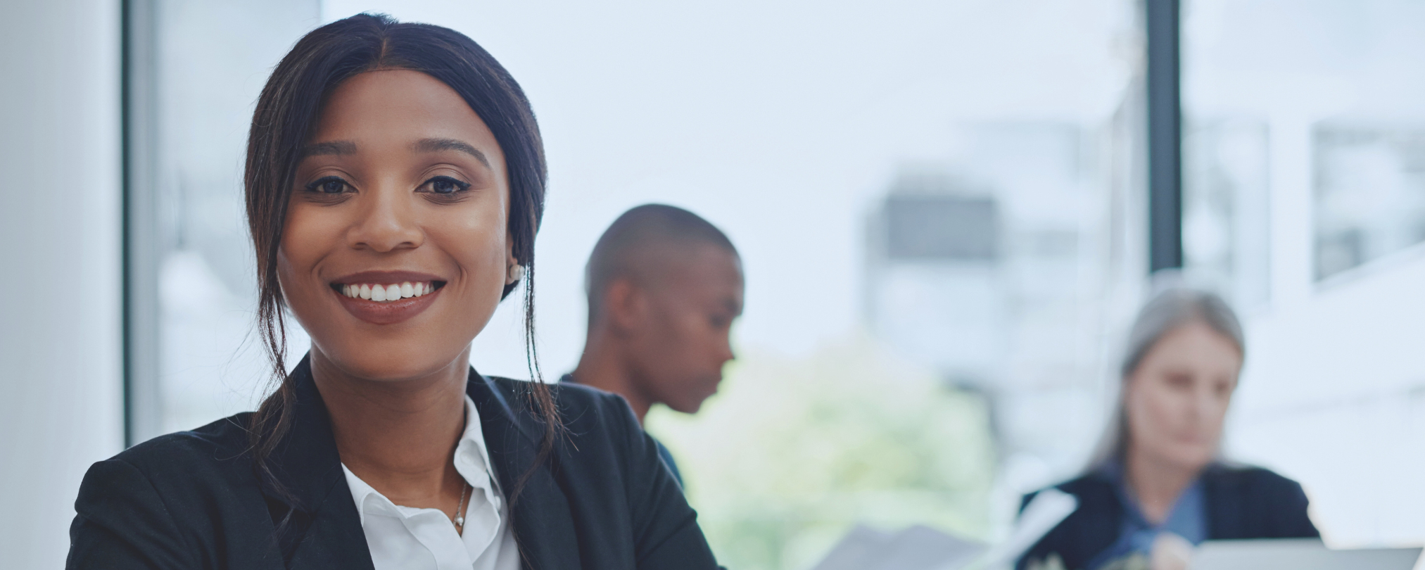 Confident professional woman smiling at the camera with colleagues working in the background.